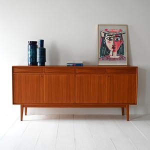 May include: A mid-century modern teak sideboard with four doors and a top shelf. The sideboard is in a light wood tone and has a minimalist design. There are three blue and green ceramic vases on the top shelf and a book titled "design*mag" on the shelf. A framed print of a colorful abstract portrait hangs on the wall above the sideboard.