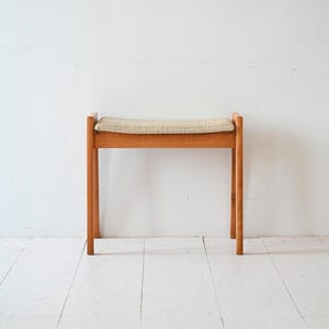 May include: A wooden stool with a light brown cushion. The stool has a simple design with four legs and a rectangular seat.