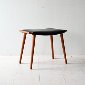 May include: A small, mid-century modern stool. It has a black seat and four wooden legs. The seat is rectangular with rounded corners. The legs are a light brown colour and angled outwards. The stool is set against a white background.