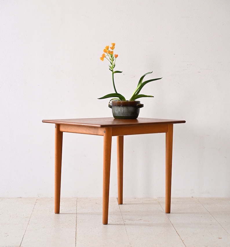 May include: A square wooden table with four legs. The table is made of light brown wood and has a smooth surface. A small potted plant with orange flowers is on the table.