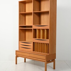 May include: A light brown wooden bookcase with multiple shelves, drawers, and compartments. The bookcase has a mid-century modern design with a raised base and vertical dividers. The drawers have black, horizontal handles.
