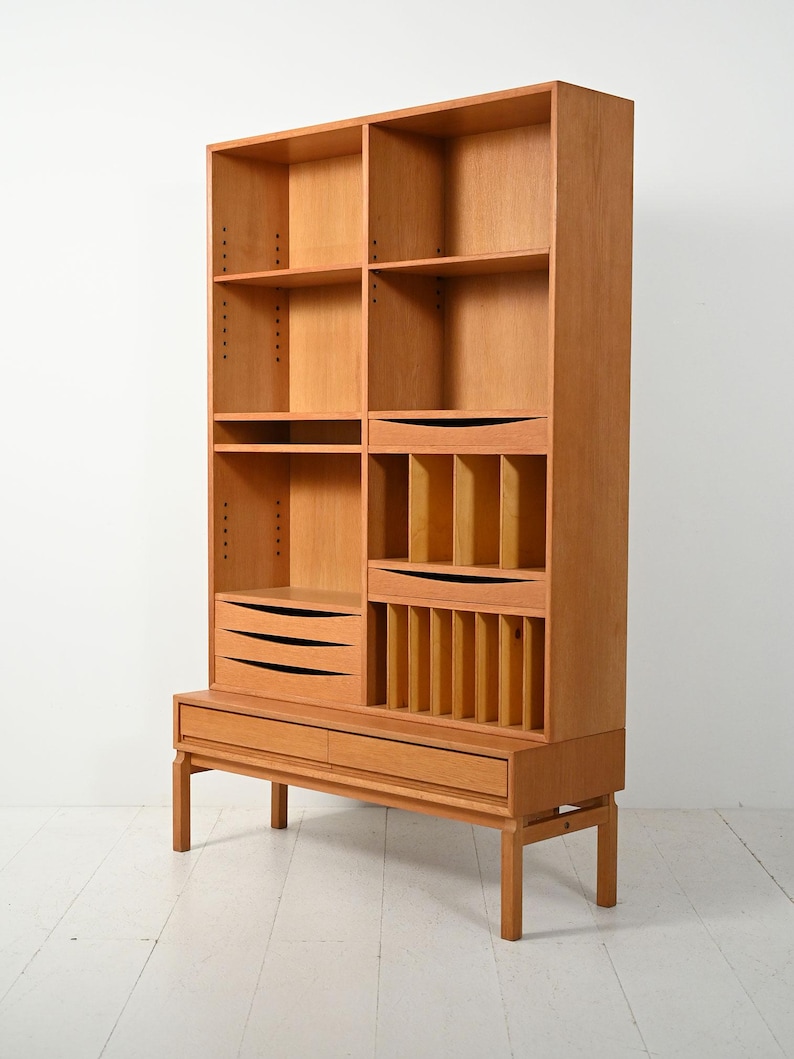 May include: A light brown wooden bookcase with multiple shelves, drawers, and compartments. The bookcase has a mid-century modern design with a raised base and vertical dividers. The drawers have black, horizontal handles.