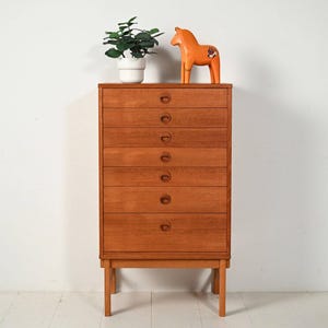 Puede incluir: Un mueble alto de madera con seis cajones y tiradores redondos. Una figura de caballo naranja y una planta en maceta están encima. El mueble es de madera marrón claro y tiene un diseño sencillo y moderno de mediados de siglo.