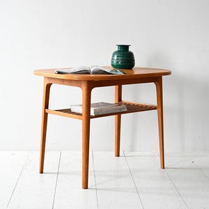May include: A mid-century modern, round side table made of light-colored wood. The table has a lower shelf and is topped with a green, textured vase and an open magazine. The table is approximately 24 inches in diameter and 20 inches tall.