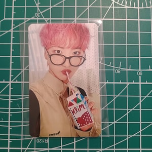 May include: A photo card featuring a person with pink hair and glasses, holding a small milk carton with a strawberry design. The card is in a clear plastic sleeve, set against a green cutting mat.