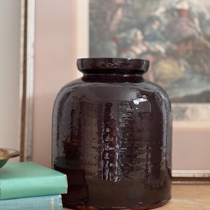 May include: A large, dark brown ceramic jar with a wide, rounded body and a slightly flared rim. The jar has a glossy finish, reflecting the light. The jar is a decorative item, suitable for home decor.