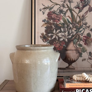 May include: A beige ceramic crock with two side handles sits on a wooden surface. Behind it is a framed floral print with a gold border. Books and a beaded necklace are also on the surface.