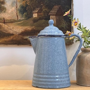 May include: A light blue enamel coffee pot with a speckled finish, a matching lid with a small knob, a curved handle, and a tapered spout. The base features three horizontal embossed rings, and the rim is accented with a thin dark blue trim.