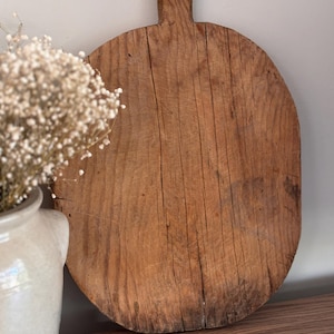 May include: A vintage, circular wooden chopping board with a handle. The board is crafted from light brown wood, displaying prominent grain and natural marks. The handle is rectangular and fixed to the top. This board is likely used for food preparation, measuring approximately 30 cm in diameter.