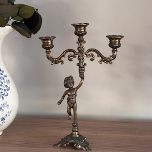 May include: A bronze-toned metal candelabra featuring a central cherub figure holding up three candle holders. The base is ornate and decorative, supporting the figure that extends upwards to the three-armed candle support structure.