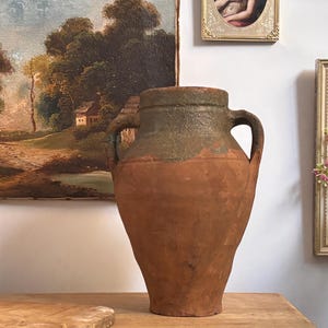 May include: A large, antique terracotta amphora with two handles. The vessel has a bulbous body and a textured, weathered surface. The top is a darker green colour. The amphora is displayed on a wooden surface, with a painting and framed art in the background.