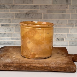 May include: A cylindrical ceramic crock with a glossy, honey-coloured glaze and a thick, rounded rim. The vessel features subtle variations in colour and texture across its surface, showing a warm, earthy tone throughout its exterior body.