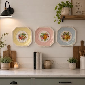 May include: Three decorative square plates in yellow, pink, and blue, each with a fruit design in the centre and a beaded edge. The plates are mounted on a white wall above a countertop with a neutral colour scheme.