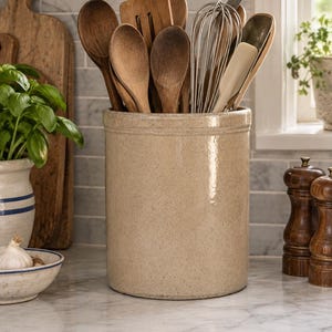 May include: A cylindrical beige ceramic crock filled with various kitchen utensils, including wooden spoons, a metal whisk, and a spatula. The container has a smooth, glossy finish and a simple, straight-sided design, holding multiple tools upright.