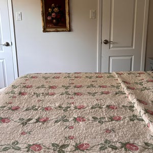 May include: A cream-coloured quilted bedspread featuring a repeating pattern of pink roses and green leaves. Above the bed, a rectangular painting in a gold frame depicts a floral arrangement of yellow, pink, and red flowers in a dark vase.