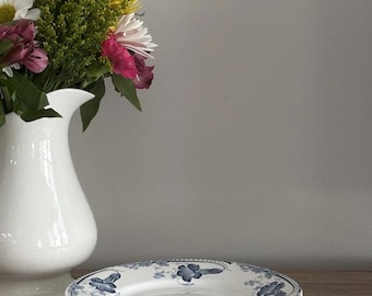 May include: A white ceramic pedestal plate with a blue floral design. The plate is on a wooden surface, next to a white vase with a bouquet of flowers. The flowers include white, pink, yellow, and green.