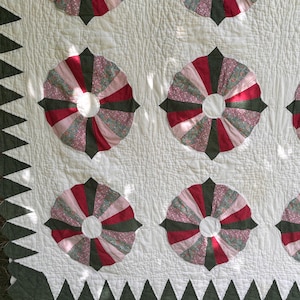 May include: A quilted textile featuring a repeating pattern of circular fan-shaped motifs in shades of dark green, deep red, and floral pink. The background is off-white with a textured surface, and the border consists of dark green triangular shapes.