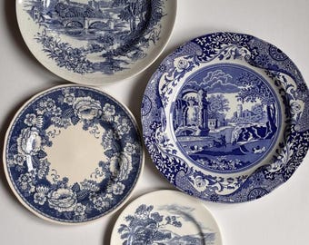 May include: Four round ceramic plates featuring blue and white transferware patterns. Three plates display detailed landscape scenes with bridges, trees, and buildings, while one plate features a central floral wreath design with roses.