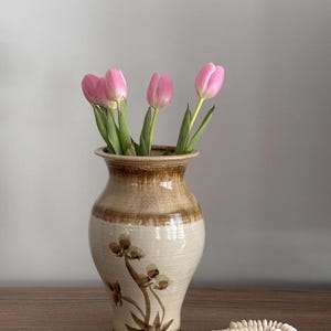 May include: A ceramic vase with a beige and brown floral design holds pink tulips. A white beaded necklace rests on a wooden surface next to the vase. The vase is approximately 20 cm tall.