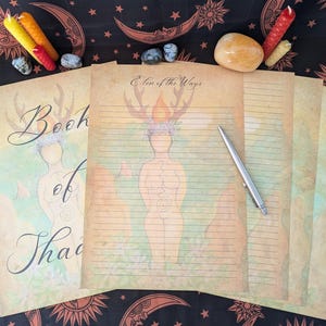 Puede incluir: Un conjunto de cuatro páginas en blanco, rayadas con un fondo de acuarela. Las páginas presentan un dibujo de una mujer con una corona de hojas y un título que dice "Elden of the Ways". Las páginas se titulan "Book of Shadows".