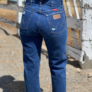 May include: Dark blue denim jeans with a classic fit. The jeans feature a high waist, back pockets, and a brown leather patch with the brand name. Distressed details add a vintage look. The jeans are worn with brown shoes.