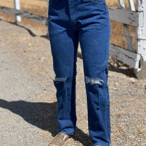 May include: Dark blue denim jeans with distressed knee details. The jeans have a classic five-pocket design and a straight leg fit. The person is wearing brown slip-on shoes.