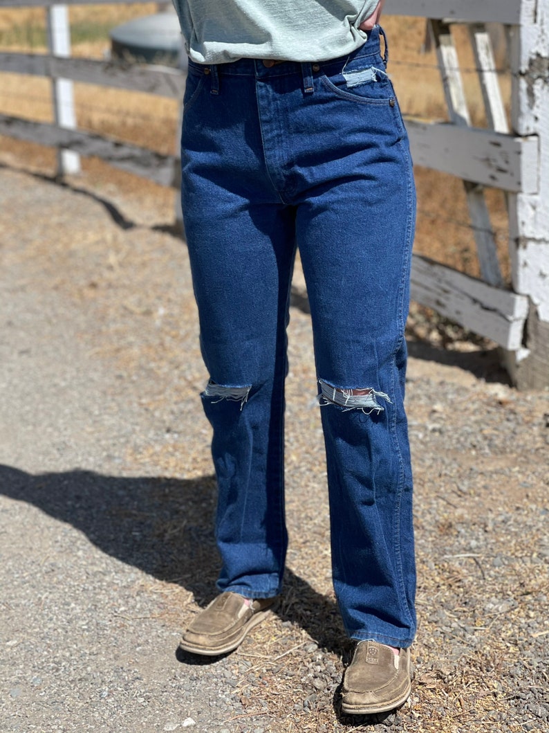 May include: Dark blue denim jeans with distressed knee details. The jeans have a classic five-pocket design and a straight leg fit. The person is wearing brown slip-on shoes.