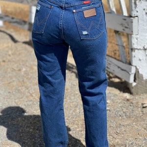 May include: Dark blue denim jeans with a classic fit. The jeans feature a distressed pocket and a brown leather patch with the brand name. The back pockets have a signature stitching design. A red tag is visible on the back pocket.