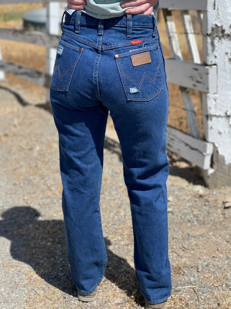 May include: Dark blue denim jeans with a classic fit. The jeans feature a distressed pocket and a brown leather patch with the brand name. The back pockets have a signature stitching design. A red tag is visible on the back pocket.