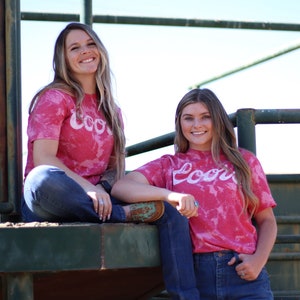 May include: Two women wearing pink tie-dye t-shirts with the word "Cool" printed on them. They are both wearing blue jeans and smiling at the camera.