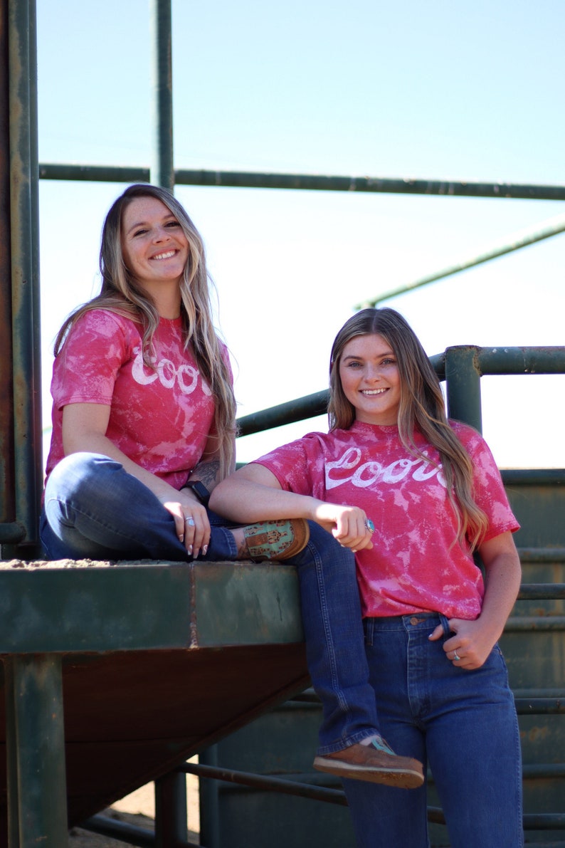 May include: Two women wearing pink tie-dye t-shirts with the word "Cool" printed on them. They are both wearing blue jeans and smiling at the camera.