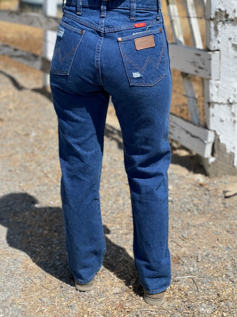 May include: Dark blue denim jeans with a classic fit. The jeans feature a high waist, back pockets, and a brown leather patch with the brand name. Distressed details add a vintage look. The jeans are worn with brown shoes.