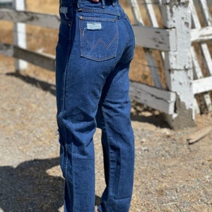 May include: Dark blue denim jeans with a classic fit. The jeans feature a Wrangler logo on the back pocket and distressed patches. The jeans have a straight leg and are worn with brown shoes.