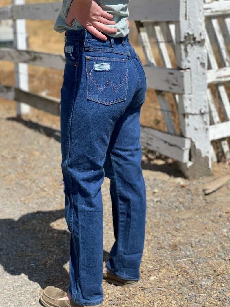 May include: Dark blue denim jeans with a classic fit. The jeans feature a Wrangler logo on the back pocket and distressed patches. The jeans have a straight leg and are worn with brown shoes.