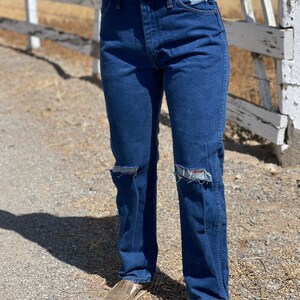 May include: Dark blue denim jeans with distressed knee details. The jeans have a classic five-pocket design and a straight leg fit. The person is wearing brown slip-on shoes. The background includes a wooden fence and a dirt path.
