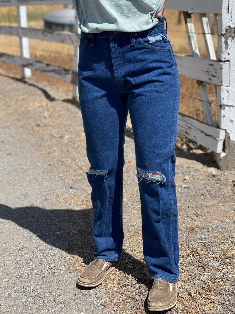 May include: Dark blue denim jeans with distressed knee details. The jeans have a classic five-pocket design and a straight leg fit. The person is wearing brown slip-on shoes. The background includes a wooden fence and a dirt path.