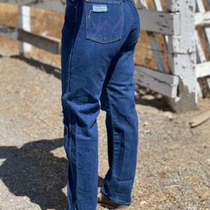 May include: Dark blue denim jeans with a classic fit. The jeans feature a distressed patch on the back pocket and the Wrangler logo. The jeans are worn with brown shoes.