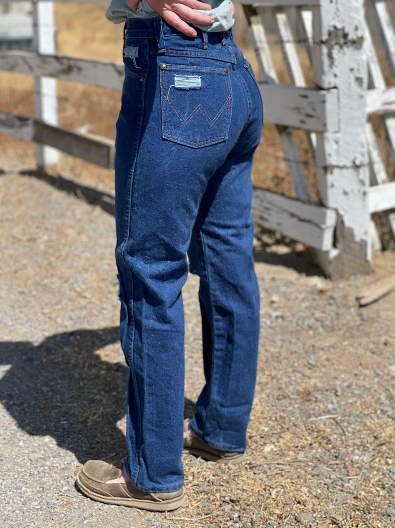 May include: Dark blue denim jeans with a classic fit. The jeans feature a distressed patch on the back pocket and the Wrangler logo. The jeans are worn with brown shoes.
