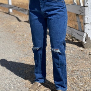 May include: Dark blue denim jeans with distressed knee details. The jeans have a classic five-pocket design and a straight leg fit. Worn with brown slip-on shoes.