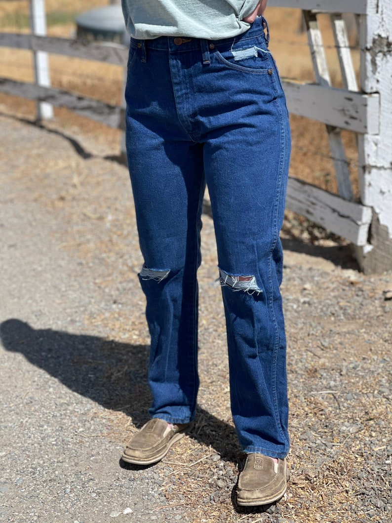 May include: Dark blue denim jeans with distressed knee details. The jeans have a classic five-pocket design and a straight leg fit. Worn with brown slip-on shoes.