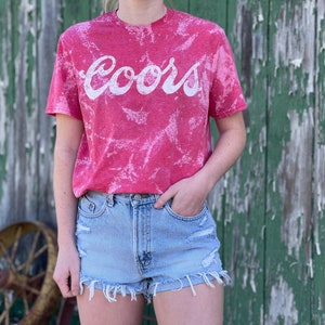 May include: A red tie-dye t-shirt with a white "Coors" logo. The shirt is worn by a person wearing light blue denim shorts.