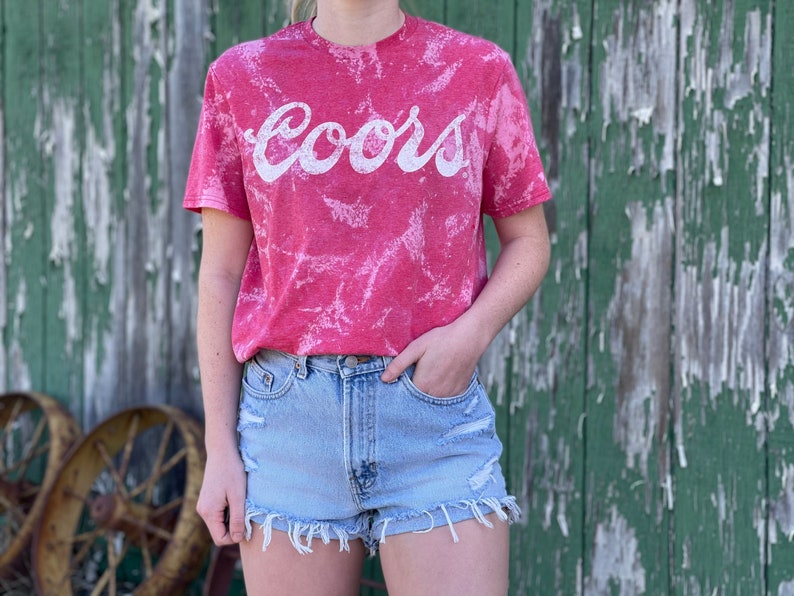 May include: A red tie-dye t-shirt with a white "Coors" logo. The shirt is worn by a person wearing light blue denim shorts.