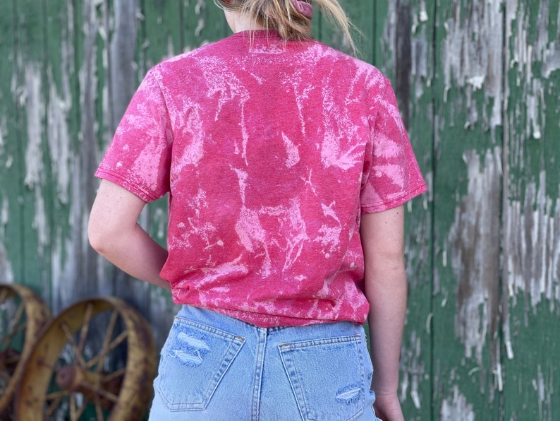 May include: A pink tie-dye t-shirt with a white bleach design. The shirt is worn by a person and is shown from the back.