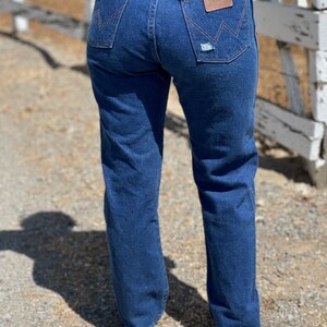 May include: Dark blue denim jeans with a classic fit, featuring distressed details on the back pockets. The jeans have a high waist and a Wrangler brand patch. The image shows the back of the jeans.