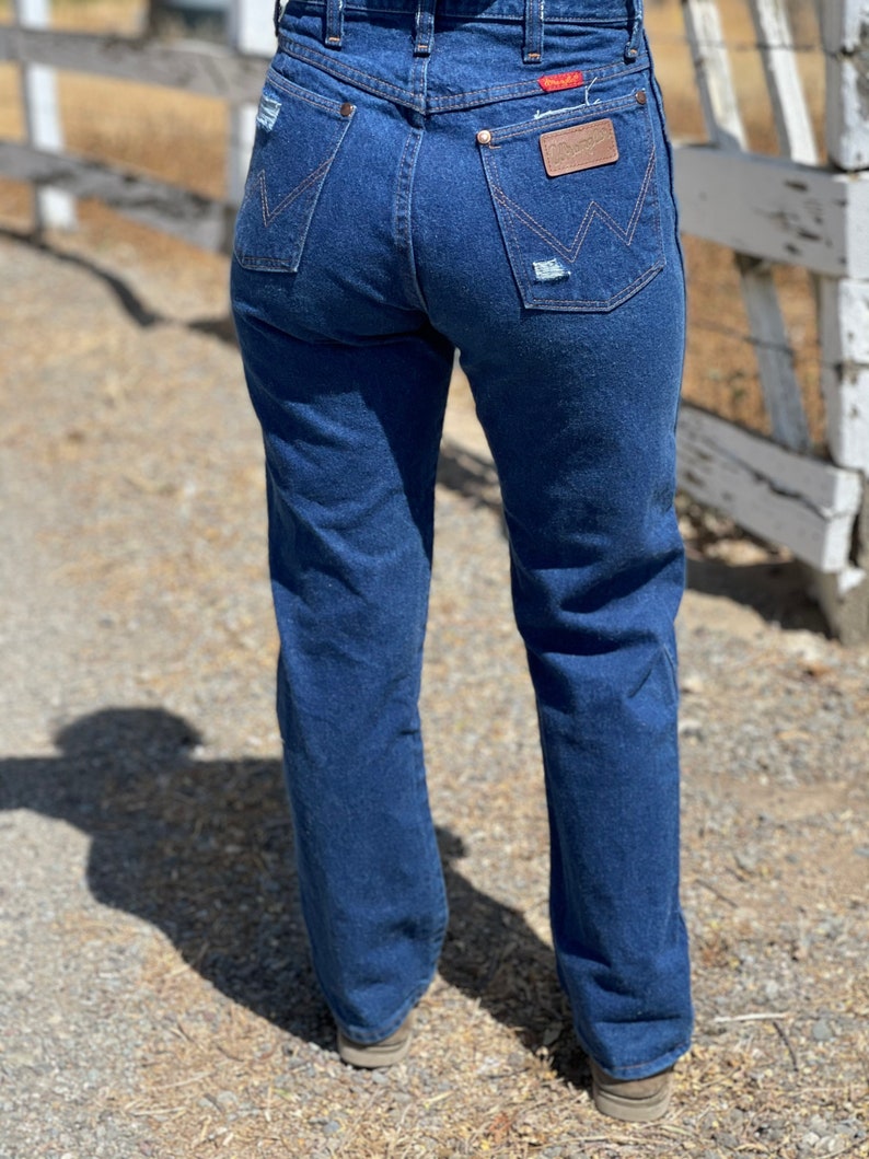 May include: Dark blue denim jeans with a classic fit, featuring distressed details on the back pockets. The jeans have a high waist and a Wrangler brand patch. The image shows the back of the jeans.