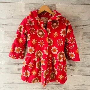 May include: A red fleece coat with a floral pattern. The coat features a collar, three buttons, and a flared skirt. The flowers are white, orange, and green. The coat is hanging on a wooden hanger.