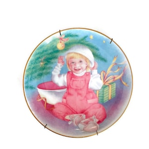 Könnte beinhalten: Dekorative Platte mit goldenem Rand, die ein Kind in rosa Latzhose und weißem Hut zeigt, mit einem Weihnachtsbaum und einem verpackten Geschenk. Der Hintergrund ist ein hellblauer Himmel.