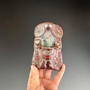 May include: A ceramic wall hanging in the shape of a stylized face with a red, white, and green glaze. The face has a hole at the top for hanging.