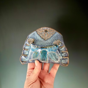 May include: A handcrafted ceramic wall pocket in shades of blue and teal, with a decorative floral design. The pocket has a rounded top with a hole for hanging and measures approximately 7 inches wide.