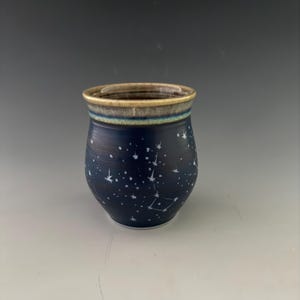 May include: A ceramic tumbler with a dark blue body featuring a white hand-painted star constellation pattern. The rim is finished with a contrasting band of light brown and teal glaze, creating a distinct border against the deep blue surface.